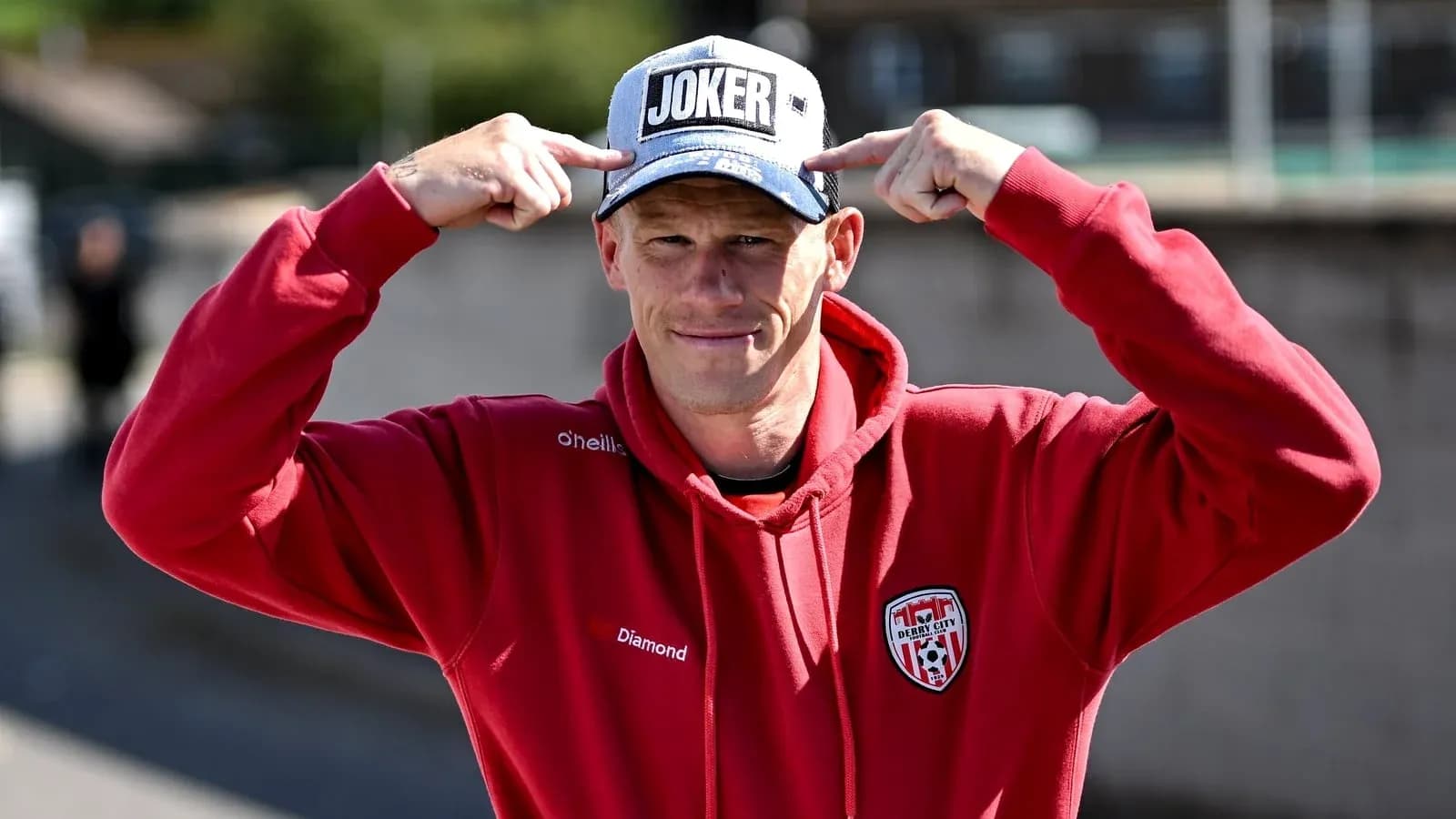 Man in red hoodie pointing at 'JOKER' cap